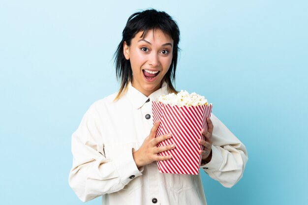 Jonge vrouw die over blauwe muur een grote emmer popcorns houdt