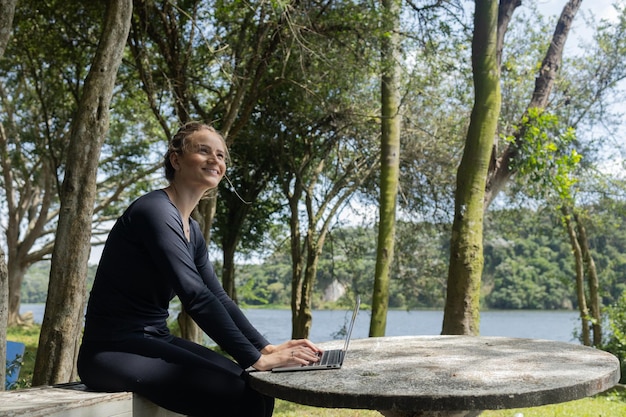 Jonge vrouw die overdag een laptop gebruikt in een groen park met mobiele telefoontechnologie