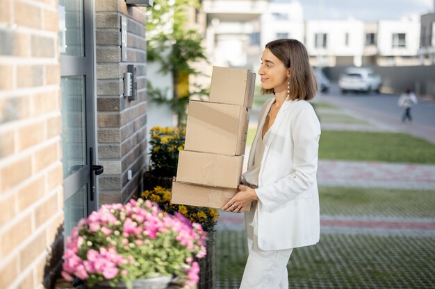 Jonge vrouw die pakjes naar huis draagt, staande met een hoop kartonnen dozen op de veranda van haar huis. Concept van online goederen kopen en thuis afleveren