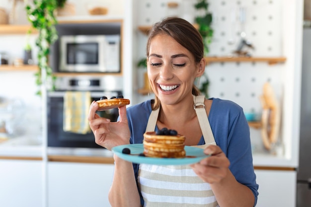 Jonge vrouw die pannenkoeken maakt in de keuken Jonge huisvrouw geniet van bosbessenpannenkoekjes als ontbijt