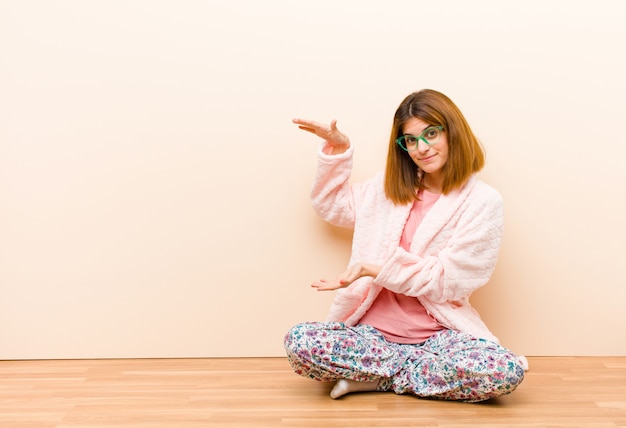 Jonge vrouw die pyjama's draagt die thuis een voorwerp met beide handen aan kant houden copyspace, een voorwerp tonen, aanbieden of adverteren