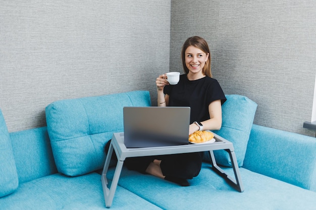 Jonge vrouw die 's ochtends koffie geniet met behulp van een laptop terwijl ze thuis op de bank zit