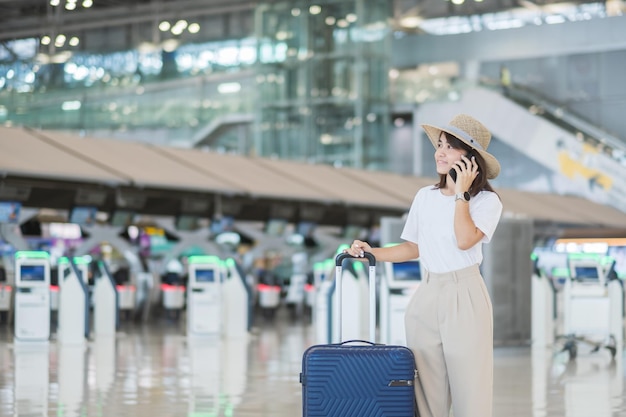 Jonge vrouw die smartphone gebruikt en bagagehandvat vasthoudt voordat ze de vluchttijd controleert op de luchthaven Transportverzekering, reis- en vakantieconcepten