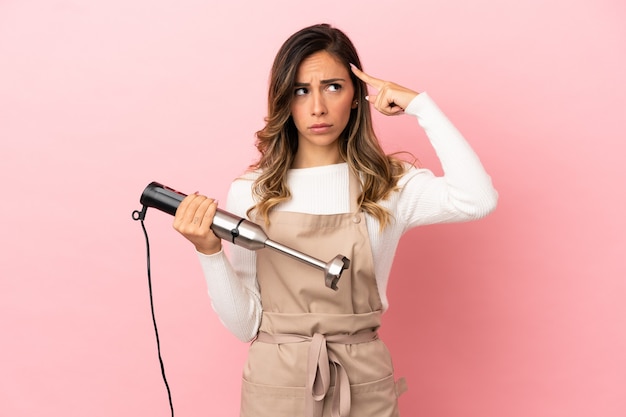 Jonge vrouw die staafmixer over geïsoleerde roze achtergrond gebruikt die twijfels heeft en denkt