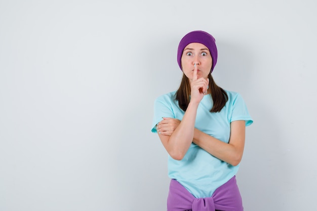 Jonge vrouw die stiltegebaar toont in blauw t-shirt, paarse muts en er bang uitziet. vooraanzicht.