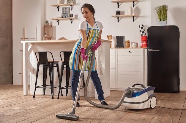 Jonge vrouw die stofzuiger in de vloer van de huiskeuken gebruikt die schoonmaaktaken en zorgvuldige karweien doet...