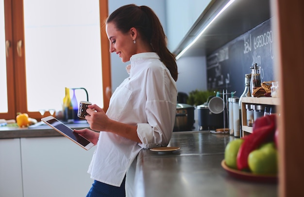 Jonge vrouw die tablet in keuken thuis gebruikt en koffie drinkt