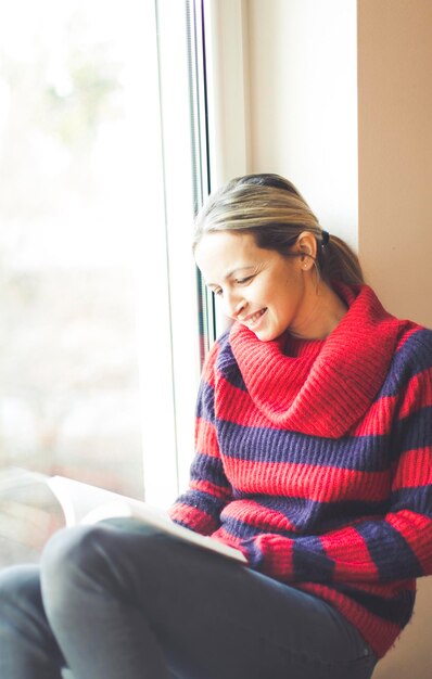 Foto jonge vrouw die thuis op de vensterbank zit en een boek leest