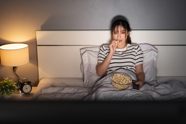 Jonge vrouw die tv kijkt en 's nachts popcorn eet op een bed