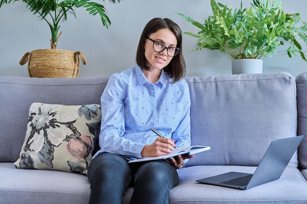 Jonge vrouw die vanuit huis werkt met behulp van laptop om aantekeningen te maken