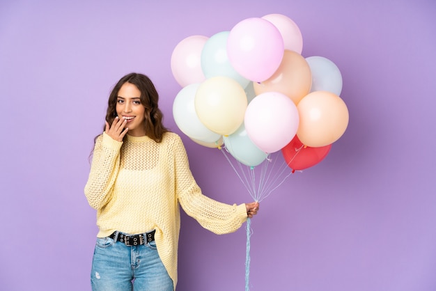 Jonge vrouw die vele ballons over geïsoleerd op purpere muur met verrassingsgelaatsuitdrukking vangt