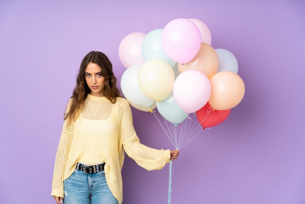 Jonge vrouw die vele ballons over geïsoleerd op purpere muur vangt die wapens gekruist houden