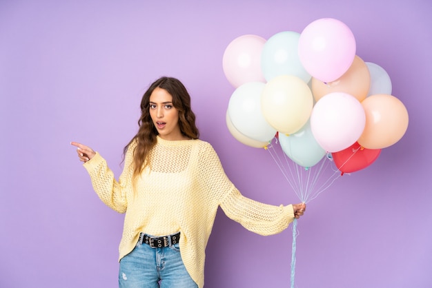 Jonge vrouw die vele ballons over geïsoleerd op purpere muur verrast verrast en vinger aan de kant richt