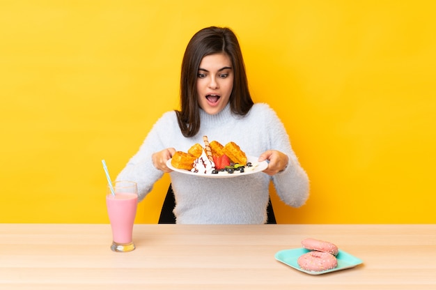 Jonge vrouw die wafels en milkshake eet in een lijst over geïsoleerde gele muur