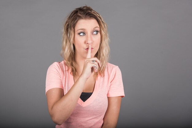 Jonge vrouw die wegkijkt met vinger op mond tegen grijze achtergrond