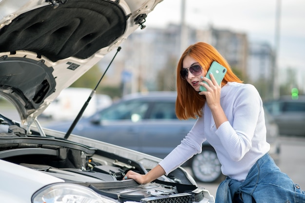 Foto jonge vrouw die zich dichtbij gebroken auto met geknalde kap bevindt die op haar mobiele telefoon spreekt terwijl het wachten op hulp.