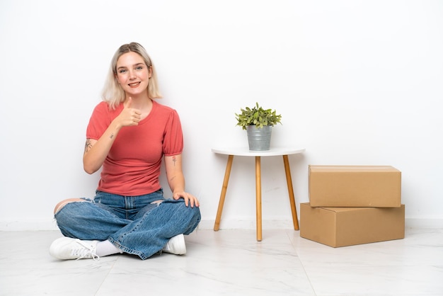 Jonge vrouw die zich in nieuw huis tussen dozen beweegt die op witte achtergrond worden geïsoleerd en een duim omhoog gebaar geeft