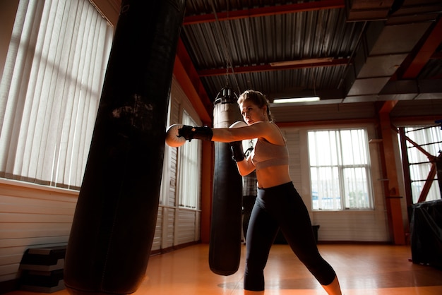 Jonge vrouw doet bokstraining in de sportschool, ze draagt bokshandschoenen en slaat een bokszak.