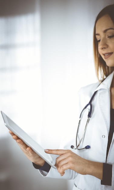 Jonge vrouw-dokter houdt een tabletcomputer in haar handen, terwijl ze in een kliniek staat. Portret van vriendelijke vrouwelijke arts met een stethoscoop op het werk. Perfecte medische dienst in een ziekenhuis. medicus