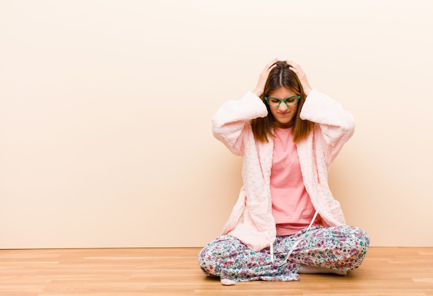 Jonge vrouw draagt pyjama's om thuis te zitten, gestrest en gefrustreerd, haar handen opheffende, moe, ongelukkig en met migraine