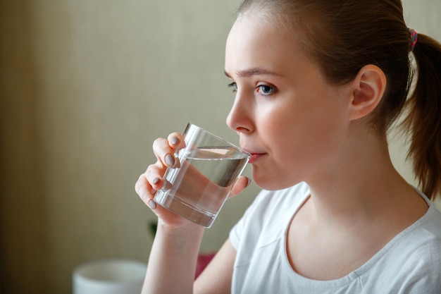 Jonge vrouw drinkt 's ochtends na het ontwaken een glas zuiver water. gelukkig tienermeisje handhaaft de waterbalans voor de gezondheid van het lichaam door een transparant kopje schoon water te drinken.