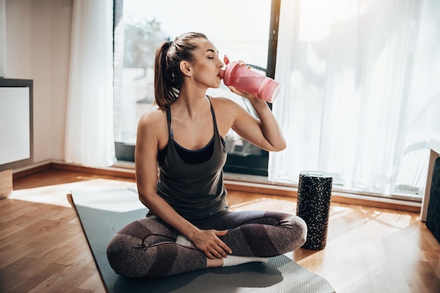 Jonge vrouw drinkt water terwijl ze zich 's ochtends thuis voorbereidt op fitnesstraining.