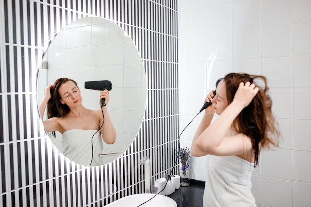 Jonge vrouw drogen en stylen van haar met föhn kapsel maken in moderne badkamer interieur Schoonheidsroutine na ochtenddouche