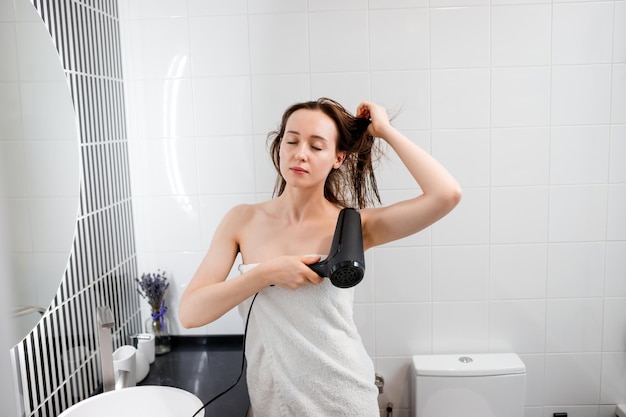 Jonge vrouw drogen en stylen van haar met föhn kapsel maken in moderne badkamer interieur Schoonheidsroutine na ochtenddouche