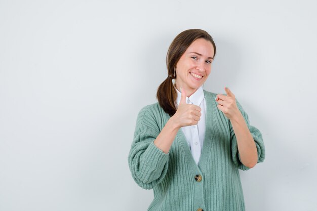 Jonge vrouw duim opdagen, naar voren wijzend in blouse, vest en vrolijk kijken. vooraanzicht.