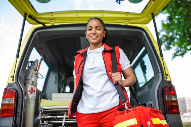 Jonge vrouw een paramedicus die aan de achterkant van een ambulance bij de open deuren staat Ze kijkt naar de camera met een zelfverzekerde uitdrukking die lacht en een medische traumatas op haar schouder draagt