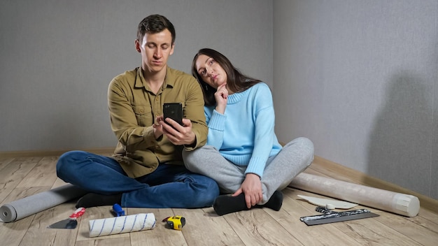 Jonge vrouw en man kiezen meubels en kamerinrichting met behulp van de telefoon en argumenteren zittend op een parketvloer tussen bouwmaterialen en gereedschappen.