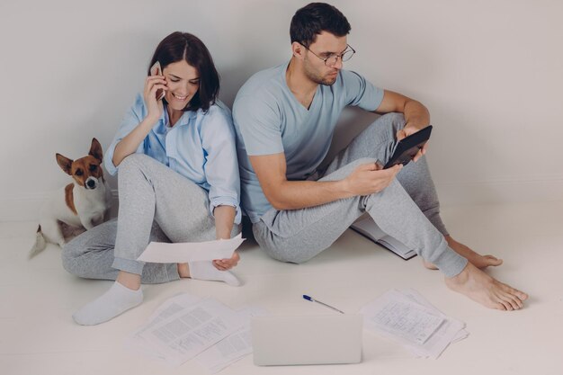 Jonge vrouw en man leun achterover tegen elkaar doen rekeningen samen maken noodzakelijke berekeningen omringd met papieren documenten zitten op de vloer in lege ruimte met hond Mensen en financiën concept