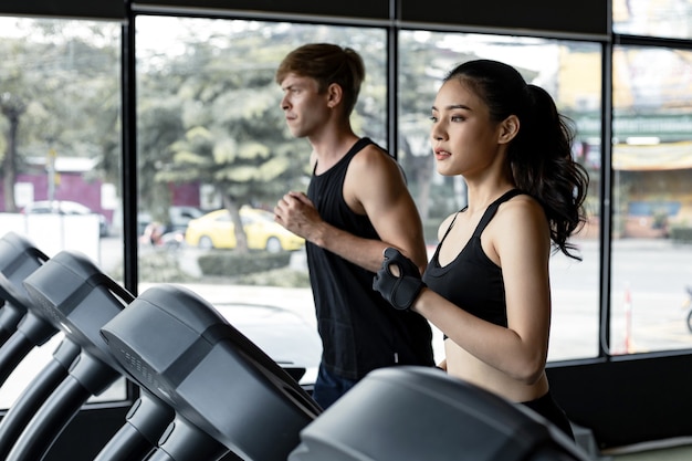 Jonge vrouw en man lopen naast elkaar op moderne elektrische loopbanden in de sportschool. vrij aziatische vrouw draait op loopband met fit jonge man op achtergrond. jong koppel op elektrische loopbanden.