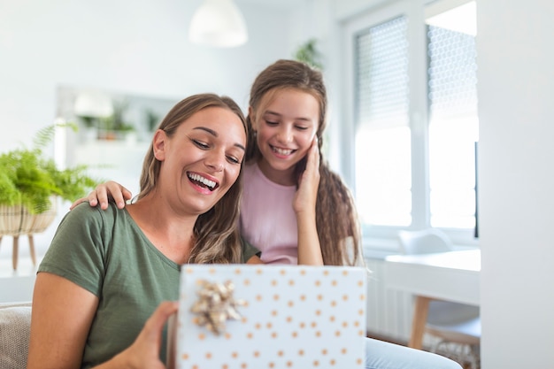 Jonge vrouw en meisje thuis vieren Moederdag zittend op de bank dochter knuffelen moeder kussen wang moeder lachen vreugdevolle holding geschenkdoos