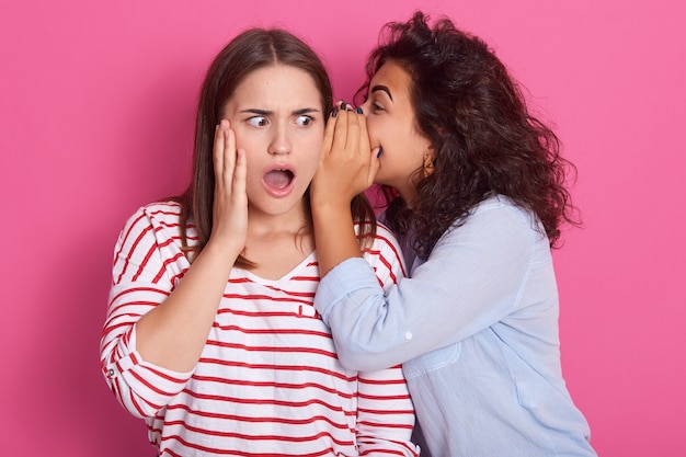 jonge vrouw fluistert schokkend nieuws tegen haar partner, brunette vrouw kleedt gestreepte casul shirt poseren