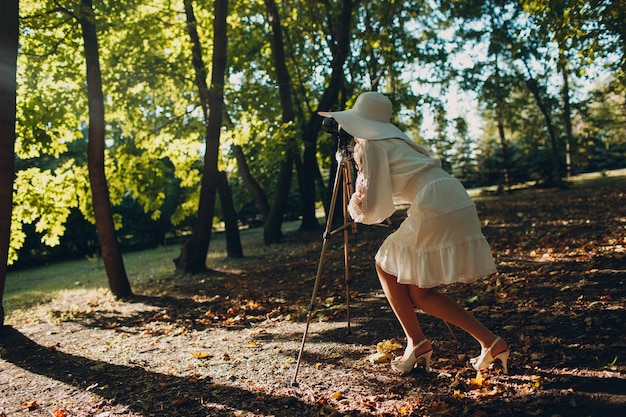 Jonge vrouw fotograaf in witte jurk en hoed met camera in herfst park.