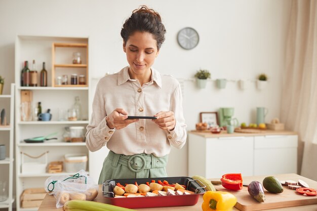 Jonge vrouw fotografeert haar speciale schotel met haar mobiele telefoon terwijl ze in de keuken staat