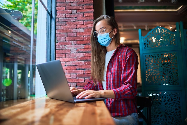 Jonge vrouw freelancer die een gezichtsmasker van de geneeskunde draagt dat op afstand werkt op een computer in een café tijdens een pandemie. sociale afstand en bescherming van de gezondheid tegen virussen op openbare plaatsen