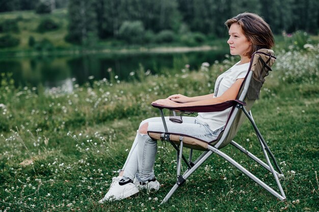 Jonge vrouw freelancer zittend op een stoel en ontspannen in de natuur in de buurt van het meer. Buitenactiviteiten in de zomer.