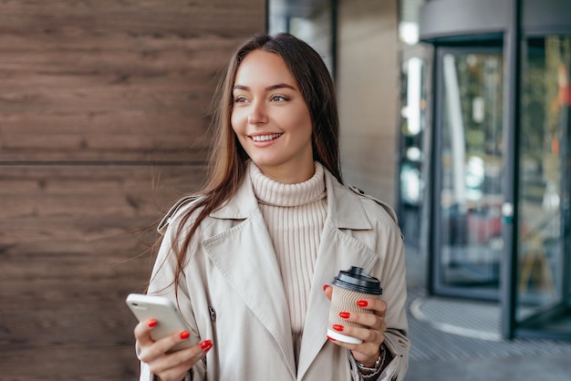 Jonge vrouw gebruikt een mobiele telefoon glimlacht en kijkt weg tegen de achtergrond van een kantoorgebouw