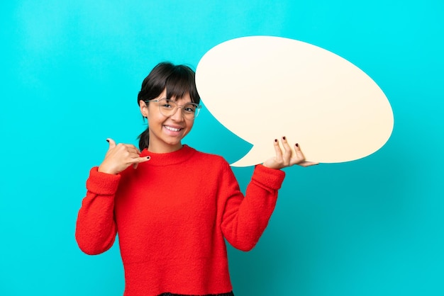 Jonge vrouw geïsoleerd op blauwe achtergrond met een lege tekstballon en telefoongebaar