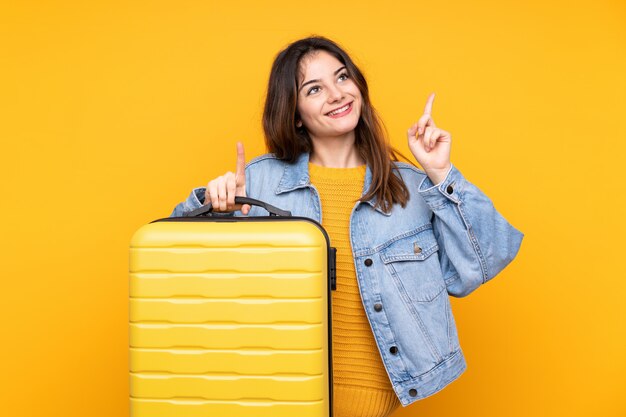 Jonge vrouw geïsoleerd op geel in vakantie met reiskoffer en omhoog