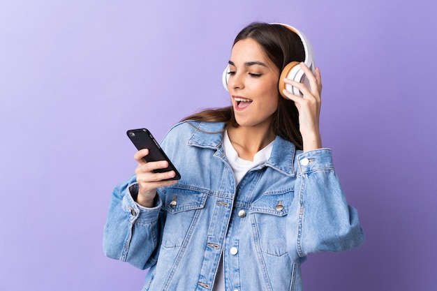 Jonge vrouw geïsoleerd op paarse muur muziek luisteren met een mobiele telefoon en zingen