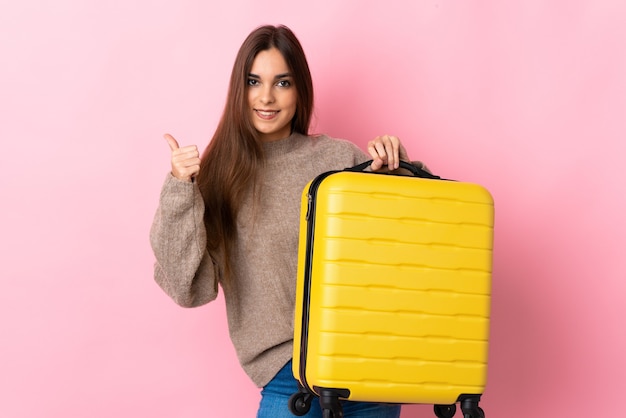 Jonge vrouw geïsoleerd op roze muur in vakantie met reiskoffer en met duim omhoog