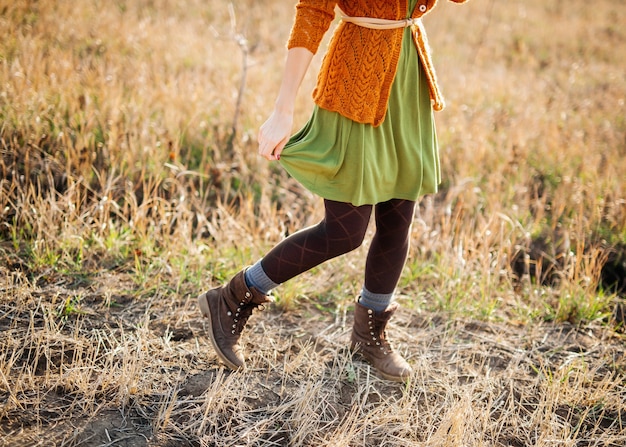 Jonge vrouw, gekleed in een jurk, gebreide cardigan en laarzen, wandelen in een veld