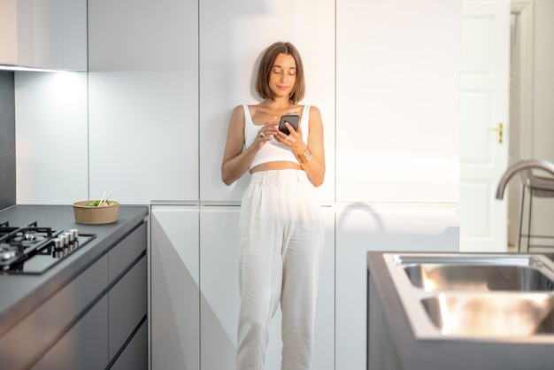 Jonge vrouw gekleed in het wit staande ontspannen met telefoon op de moderne keuken thuis. Smart home en modern woonconcept