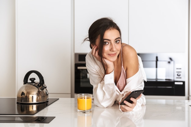 jonge vrouw, gekleed in zijden kleding sap drinken in de keuken, en het gebruik van zwarte mobiele telefoon met een glimlach