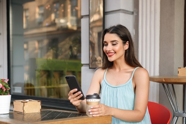 jonge vrouw gelukkig zitten in café buitenshuis tijdens het gebruik van mobiele telefoon en koffie drinken