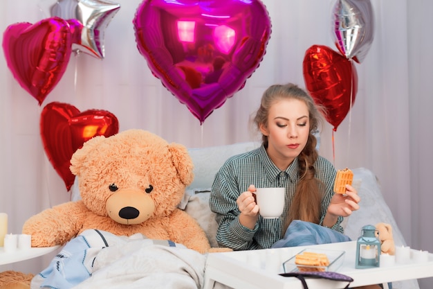 Jonge vrouw geniet van ontbijt op bed met speelgoed beer en ballonnen