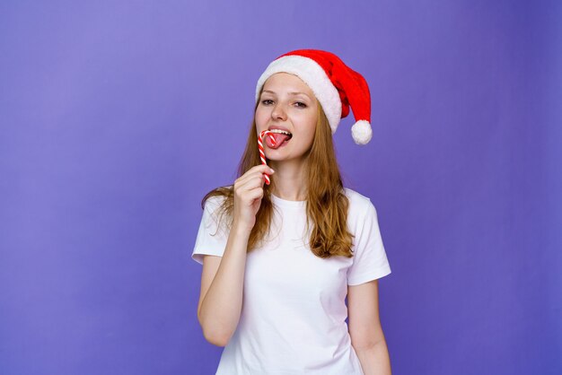 Jonge vrouw genieten van snoep in kerstmuts en witte t-shirt op lila achtergrond meisje kauwen op een kerst...